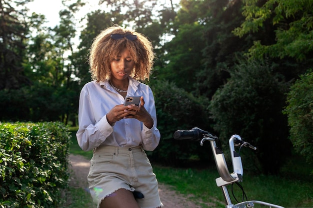 african american curly girl sits on bike in the park in summer and uses smartphone