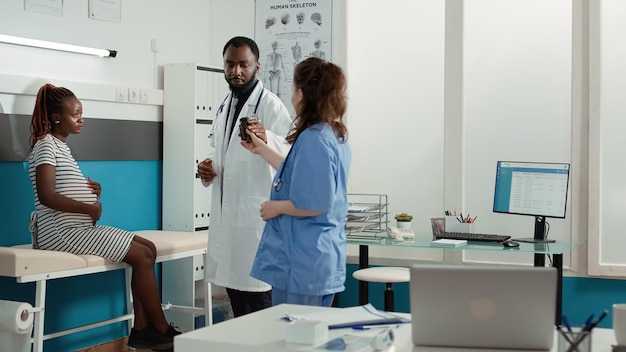 African american doctor giving bottle of pills to pregnant woman in health care service cabinet. Female patient with pregnancy belly receiving prescription medicine from general practitioner.