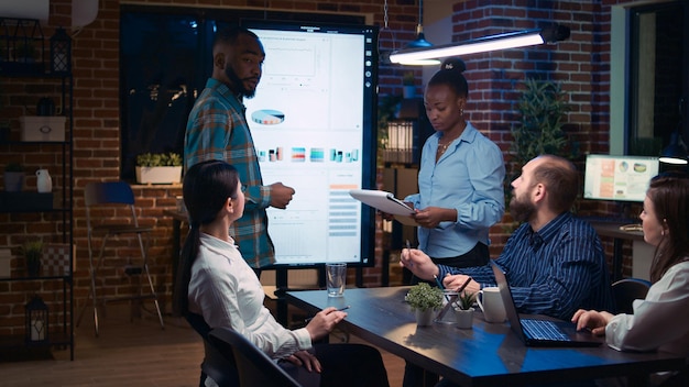 African american employees presenting company revenue statistics visualization in business meeting, people listening to presentation. Office workers in coworking space boardroom