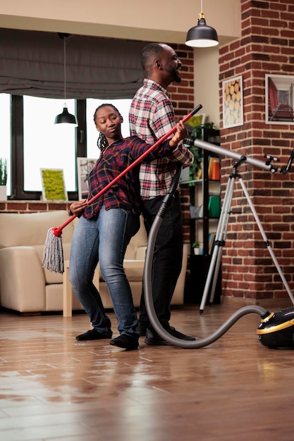 African american family doing housework in urban apartment, playing musician with mop and vacuum cleaner. Enthusiastic married couple feeling joy while doing household chores.