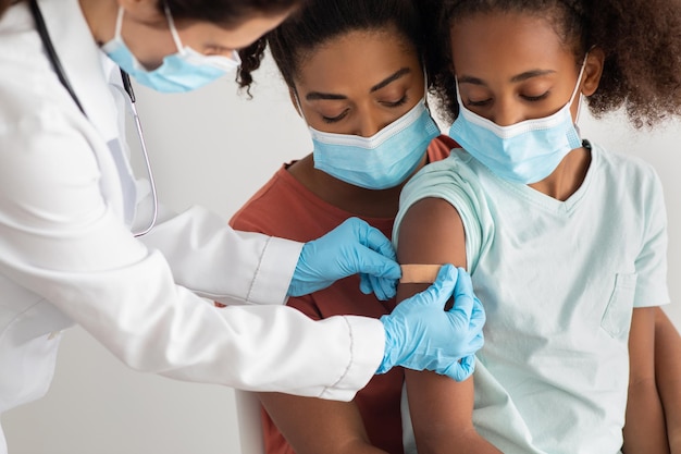 African american family young mother and teen daughter making vaccination at clinic, wearing medical face masks. Nurse putting band on shoulder after injection against COVID-19 for black girl