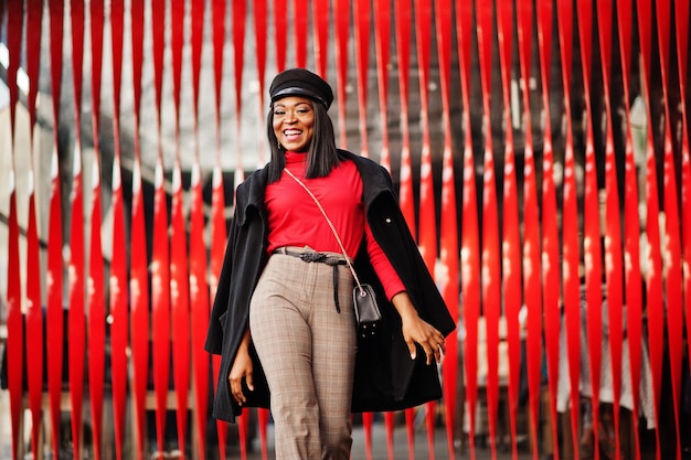 African american fashion girl in coat and newsboy cap posed at street against red background