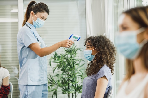 African American female teenager with a face mask having her temperature checked before coronavirus vaccine.