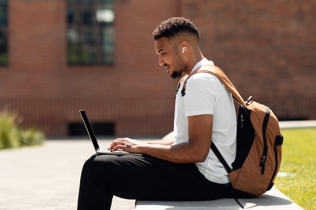 African american freelance man sitting outdoors in park typing on laptop while working online with