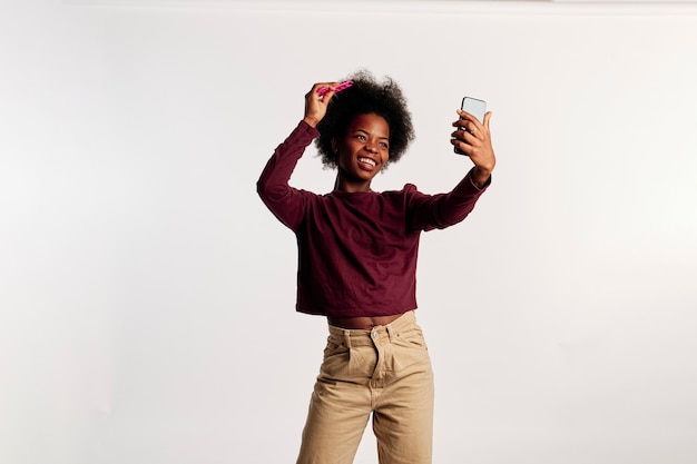 African American girl in brown sweater poses while looking at her phone and combing her hair with a pink brush.