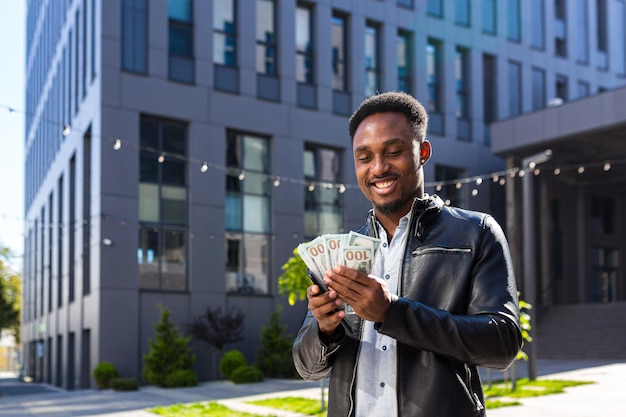 African american guy standing on street modern building rejoices show earnings winning at betting cash money with mobile phone in hands. Happy glad black man outdoors with smartphone and pack dollars