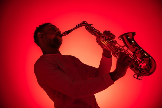 African American handsome jazz musician playing the saxophone in the studio on a neon background. Music concept. Young joyful attractive guy improvising. Close-up retro portrait.