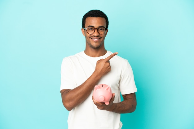 African American handsome man holding a piggybank over isolated blue background pointing to the side to present a product