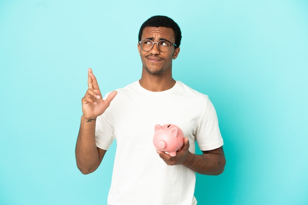 African American handsome man holding a piggybank over isolated blue background with fingers crossing and wishing the best