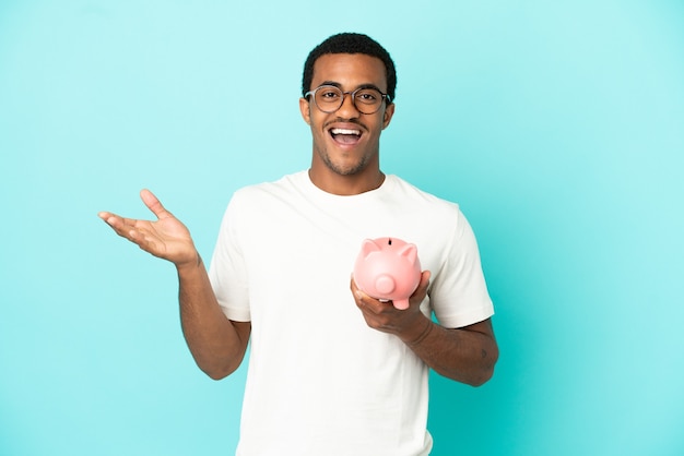 African American handsome man holding a piggybank over isolated blue background with shocked facial expression