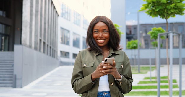 African American joyful young stylish woman tapping or scrolling on smartphone and standing at city street. Beautiful happy female texting message on mobile phone and smiling. Outside.