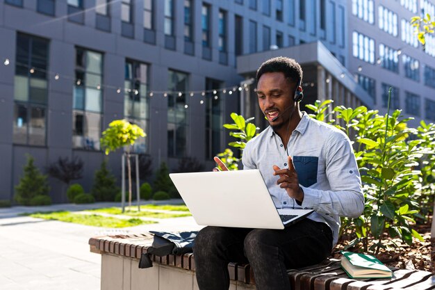 African american male freelancer working remotely online with headset and laptop. video call conference or distances meeting. Male in city park on a bench a modern urban background, outside, outdoors