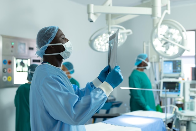 African american male surgeon studying x-ray in operating theatre, with colleagues behind