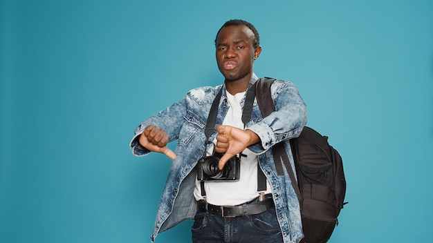 African american man doing thumbs down sign, taking pictures with dslr camera as photographer. Leaving on holiday trip with backpack to capture photos, showing disapproval and disagrement.