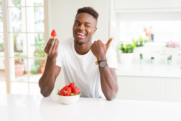 African american man eating fresh healthy strawberries pointing and showing with thumb up to the side with happy face smiling