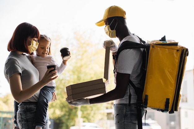 African american man in face mask works on delivery of pizza Young woman holding little daughter