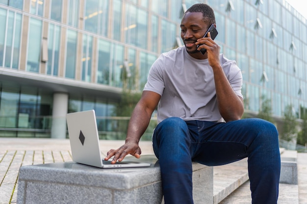 African american man freelancer using laptop talking on phone on urban street in city business man