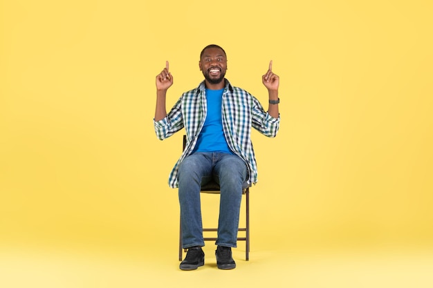 African American Man Pointing Fingers Upward Sitting Over Yellow Background