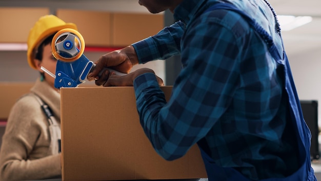 African american man putting adhesive tape on boxes, preparing order with warehouse merchandise in storage room. Male employee sealing off packages with products, delivery service.