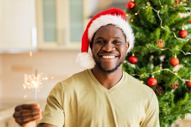 African american man in red Santa hat holding sparkler near christmas tree at home