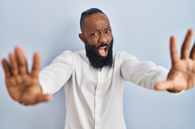 African american man standing over blue background doing stop gesture with hands palms, angry and frustration expression