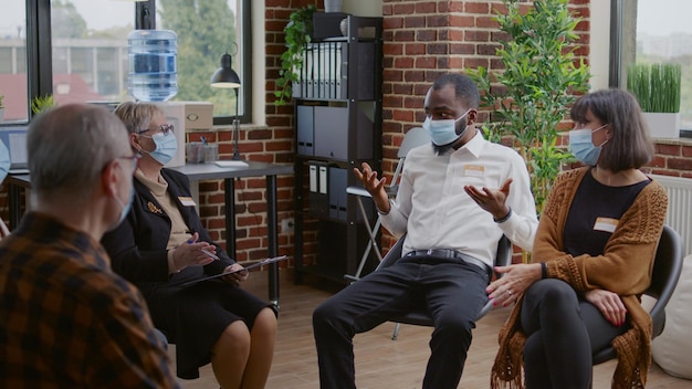 African american man talking to group of people at aa therapy meeting about addiction during pandemic. Person with face mask sharing mental health issues with patients and psychotherapist.