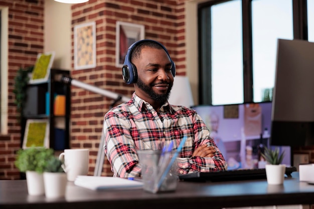 African american man using headphones and working remotely on computer at home desk. Listening to music or podcast show online on social media internet, attending class lesson webinar.