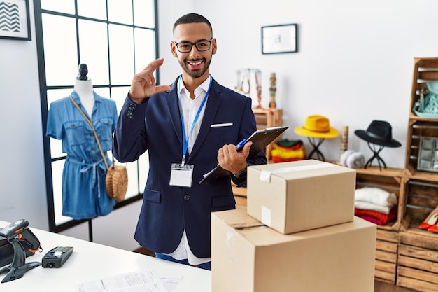 African american man working as manager at retail boutique smiling and confident gesturing with hand doing small size sign with fingers looking and the camera. measure concept.