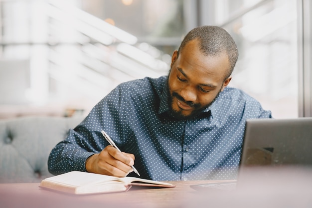 African-american man working behind a laptop and writing in a notebook. Man with beard sitting in a cafe.