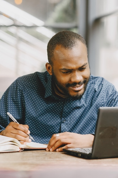 African-american man working behind a laptop and writing in a notebook. Man with beard sitting in a cafe.