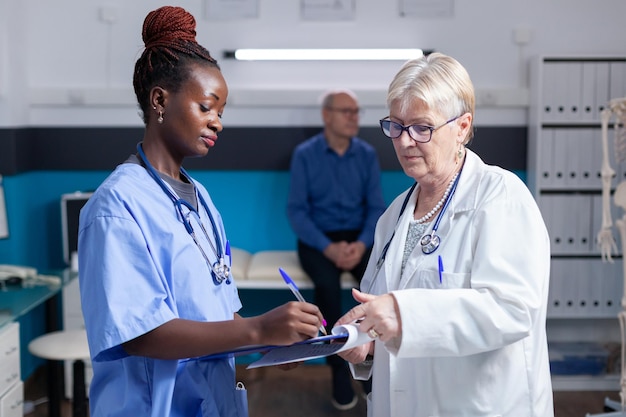 African american nurse signing checkup papers while doctor holding clipboard in cabinet. Medical assistant and physician looking at healthcare information and data on files at checkup visit