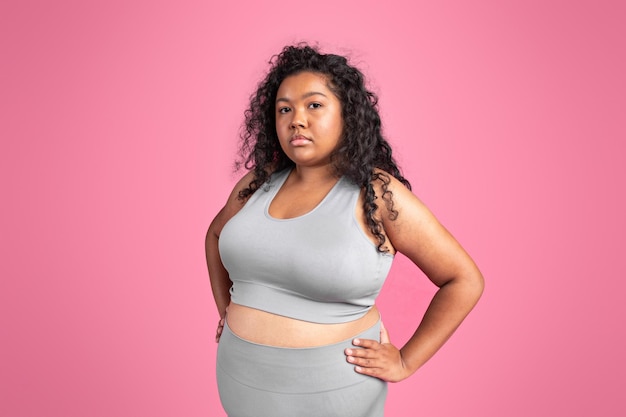 African american oversize lady in sportswear standing with hands on hips posing over pink background