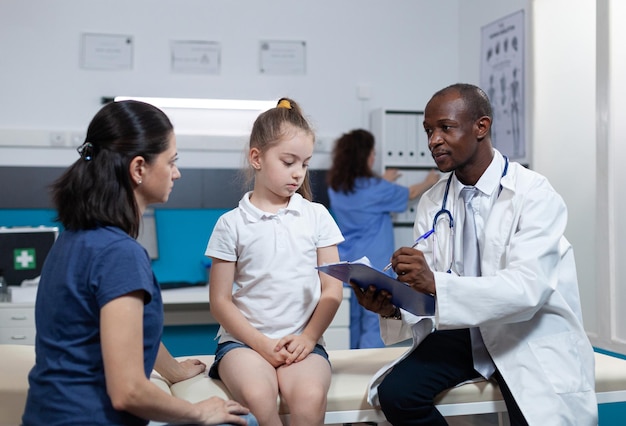 African american pediatrician doctor writing disease expertise of child on clipboard discussing medical treatment during clinical appointment in hospital office. Therapist man analyzing ill symptoms