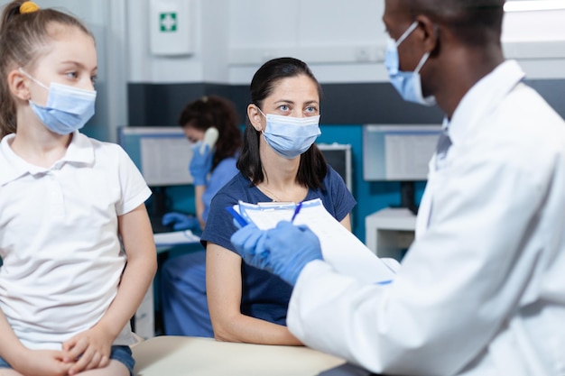 African american pediatrician doctor writing sickness symptoms on clipboard discussing healthcare treatment with family during clinical appointment in hospital office. Medical team with face mask