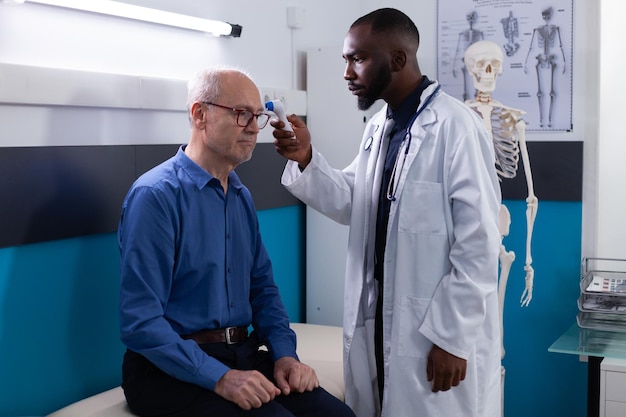 African american physician doctor putting infrared thermometer on senior man patient foredhead measuring temperature hospital office. Therpist explaining flu symtptoms during medical consultation in