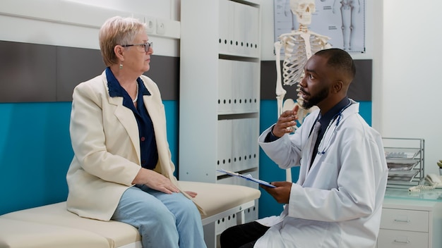 African american physician doing consultation with old woman in doctor office, explaining medical diagnosis at checkup visit appointment. Medic and patient talking about healthcare support.