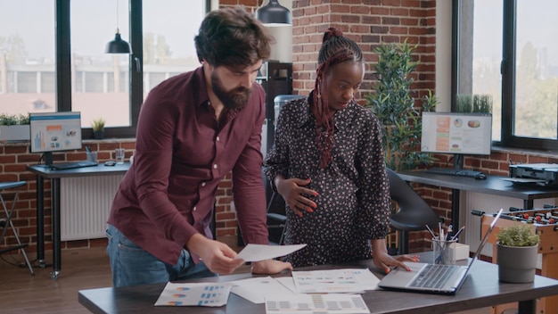 African american pregnant woman working on project planning with man at office. Business people doing teamwork with papers, talking about development. Pregnant employee expecting child