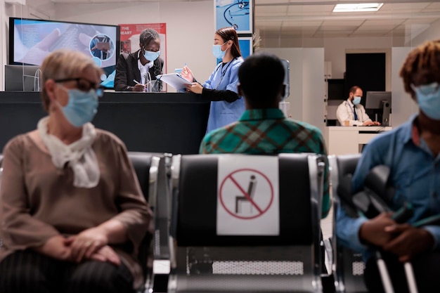 African american receptionist talking to asian nurse at reception desk in hospital lobby, chatting about healthcare service and medical appointment. Facility staff during covid 19 epidemic.