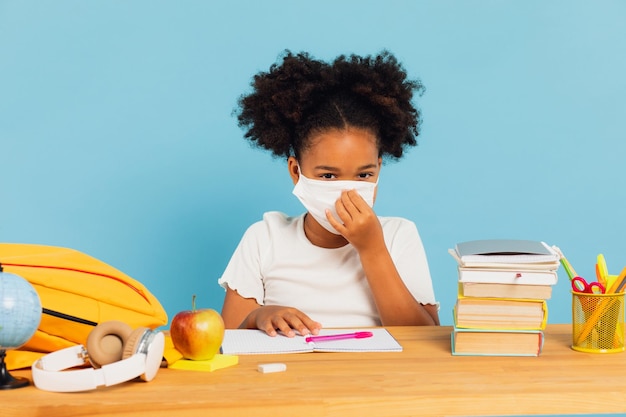 African American schoolgirl sitting at a desk in class and wearing a mask on a blue background Back to school and covid19 concept