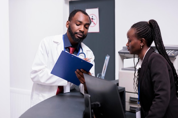 African american staff working on medical reports at hospital reception desk, analyzing patients appointments on papers. Doctor and receptionist using checkup forms to help people with disease.