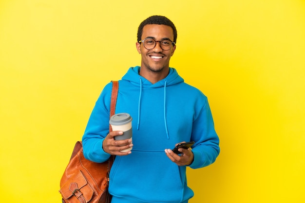 African American student man over isolated yellow wall holding coffee to take away and a mobile