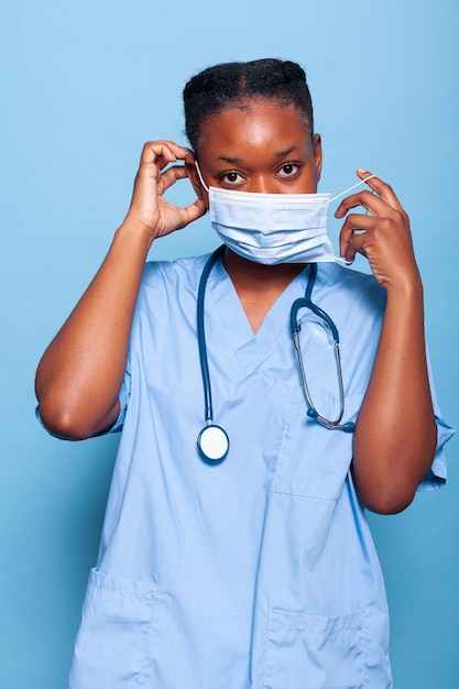 African american therapist assistant putting protection face mask to prevent infection with coronavirus while working at medical treatment in studio with blue background. Health care service