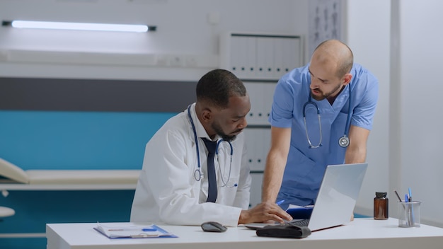 African american therapist doctor sitting at desk typing medical expertise while discussing medication treatment with man nurse analyzing sickness report. Multi-ethnic team working in hospital office