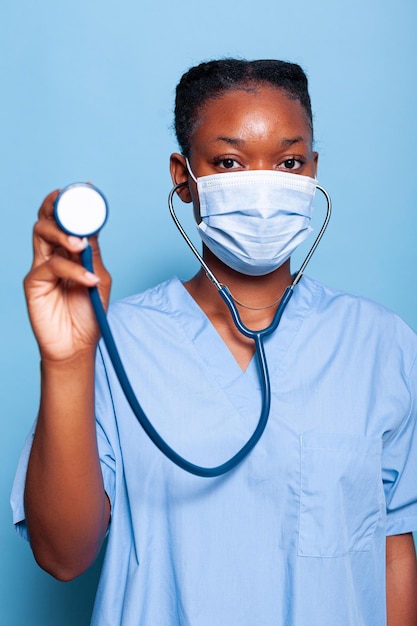 African american therapist nurse with face mask against coronavirus points medical stethoscope at camera as if she listening to patient working in studio with blue background. Medicine concept