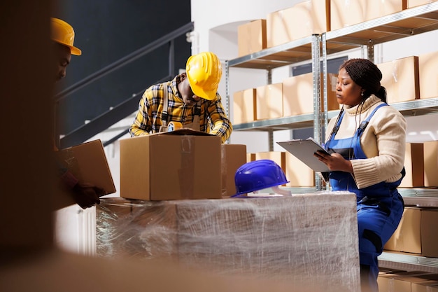 African american warehouse manager controlling assistant packing cardboard box. Supervisor holding clipboard with instruction and watching storehouse worker sealing cardboard box with adhesive