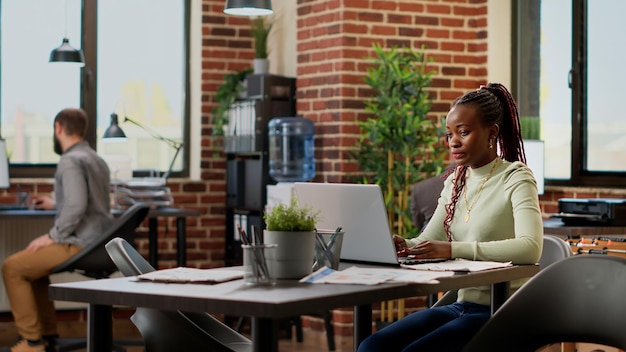 African american woman analyzing charts on papers and laptop, working on business project with data research to do report. Female employee using networking presentation online.