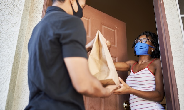 african american woman opening door to receive take out order while wearing mask