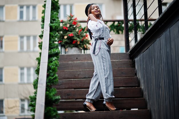 African american woman in overalls and beret posed in outdoor terrace with christmas decorations garland.