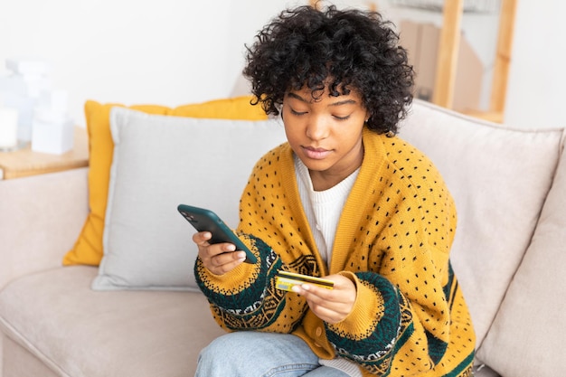 African american woman shopping online holding smartphone paying with gold credit card girl sitting