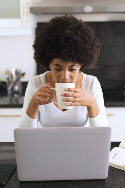 African american woman sitting in kitchen drinking coffee and using laptop